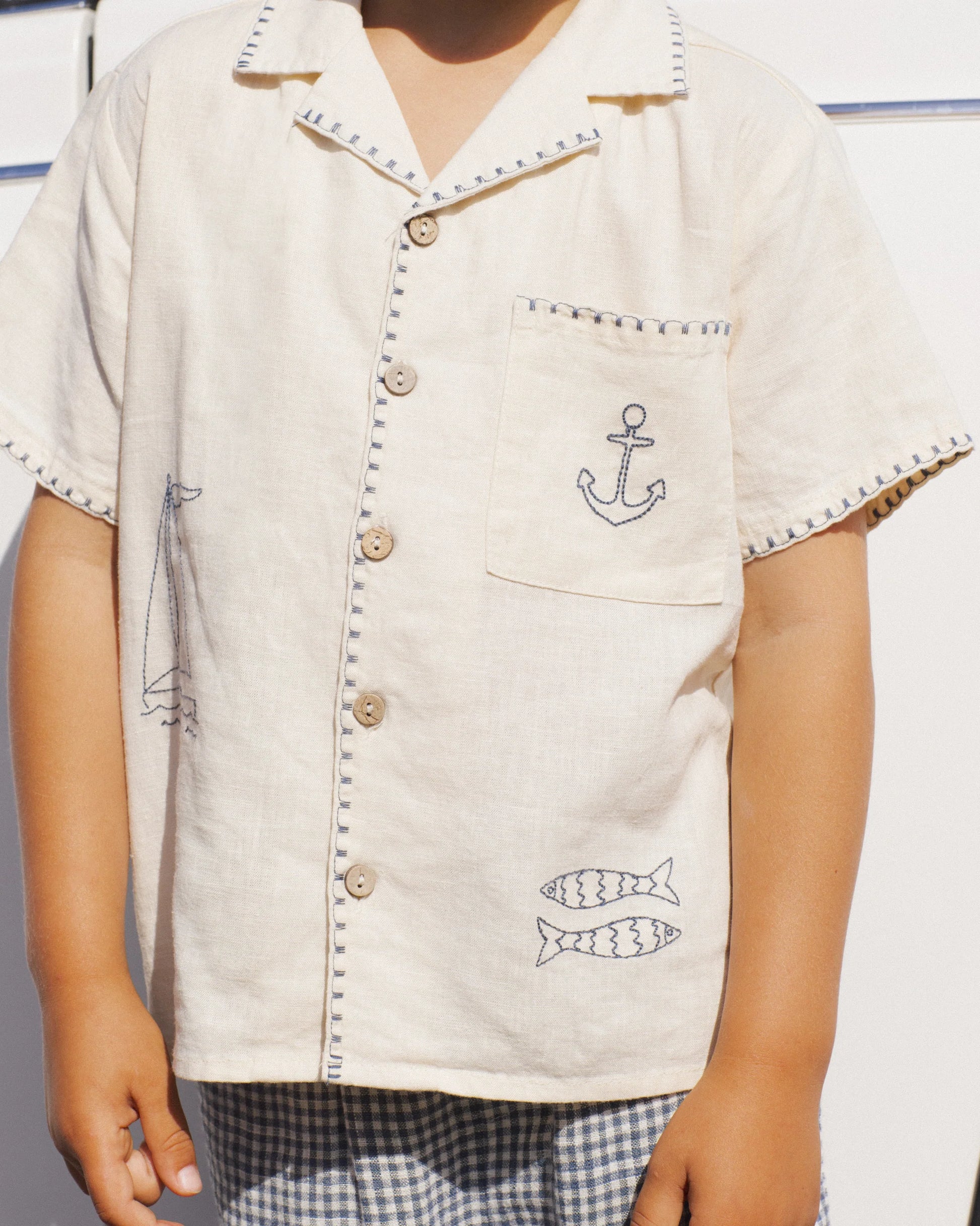 A child wears the Rylee & Cru Boys' Lapel Collar Shirt, a short-sleeved cream top with anchor, boat, and fish embroidery, paired with checkered shorts, standing against a white background.