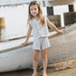 A young girl in the Rylee & Cru Girls' Knit Vest Set stands barefoot on sandy shores by a boat and fishing net, with a wooden pier in the background.