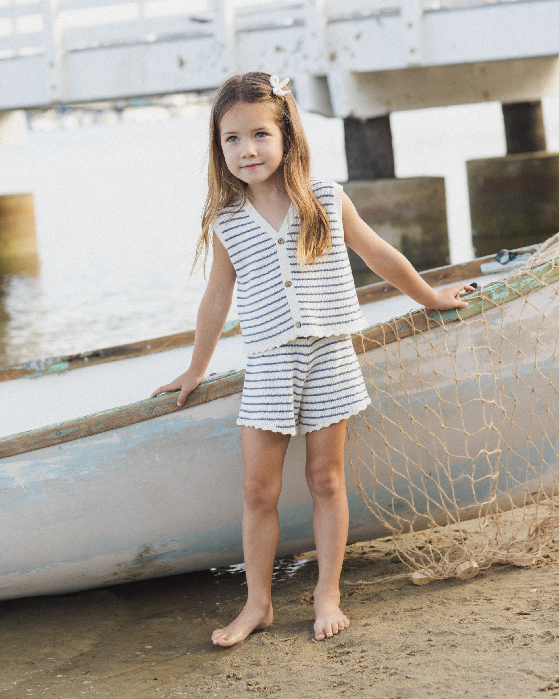 A young girl in the Rylee & Cru Girls' Knit Vest Set stands barefoot on sandy shores by a boat and fishing net, with a wooden pier in the background.