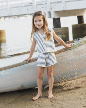 A young girl in the Rylee & Cru Girls' Knit Vest Set stands barefoot on sandy shores by a boat and fishing net, with a wooden pier in the background.