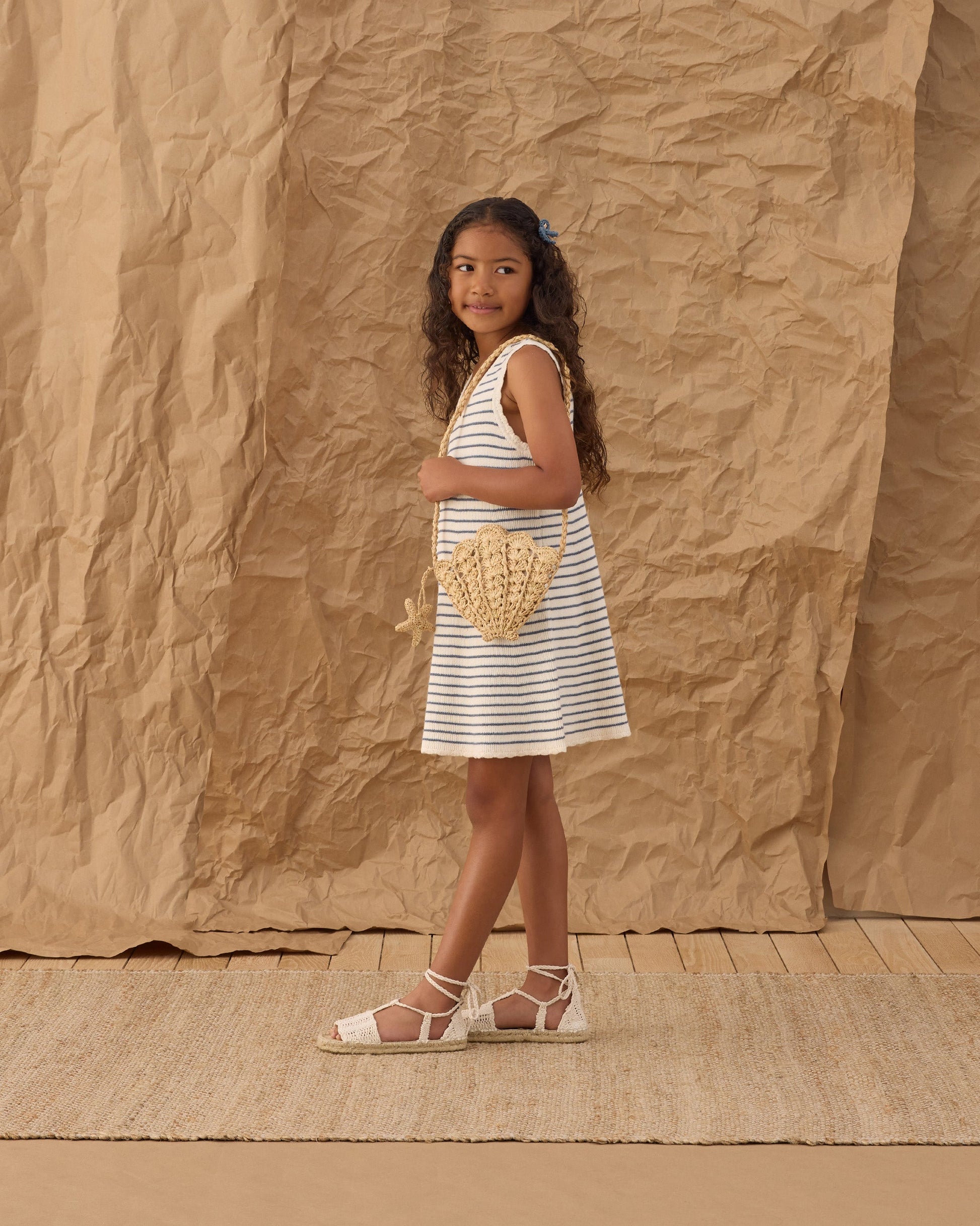 Young girl in the Rylee & Cru Girls' Crochet Tank Mini Dress, holding a straw purse and standing on a mat with a crumpled paper backdrop.