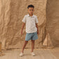 A young boy stands on a woven rug, wearing a navy stripe short-sleeve shirt and the Dawson Short from Rylee & Cru. A small wicker sailboat decorates the crumpled brown paper background.