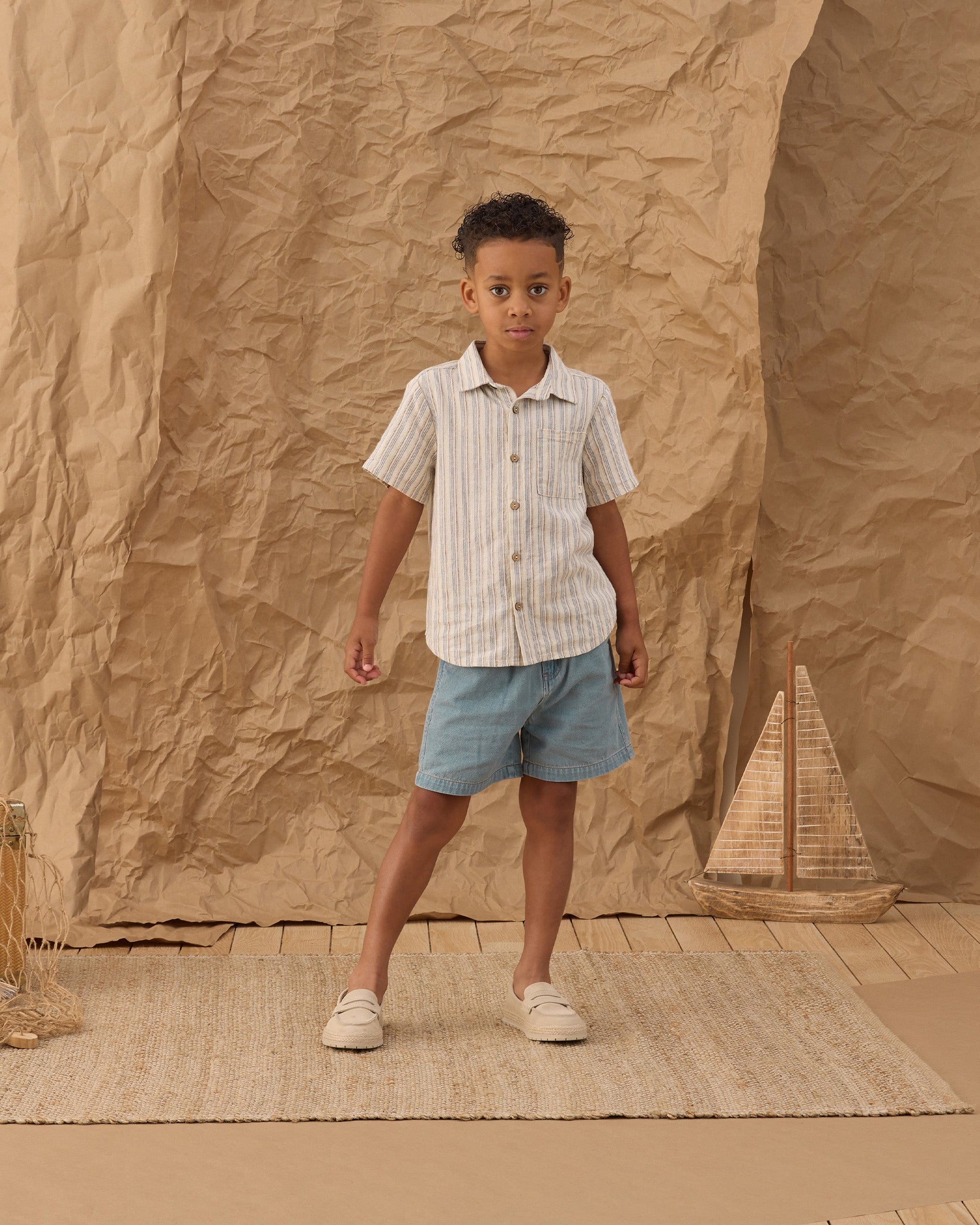 A young boy stands on a woven rug, wearing a navy stripe short-sleeve shirt and the Dawson Short from Rylee & Cru. A small wicker sailboat decorates the crumpled brown paper background.