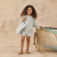 A young child with curly hair stands indoors, dressed in a light blue textured top, matching shorts, and sandals, holding the Rylee & Cru Beach Bag—an everyday accessory—beside a weathered boat adorned with netting.