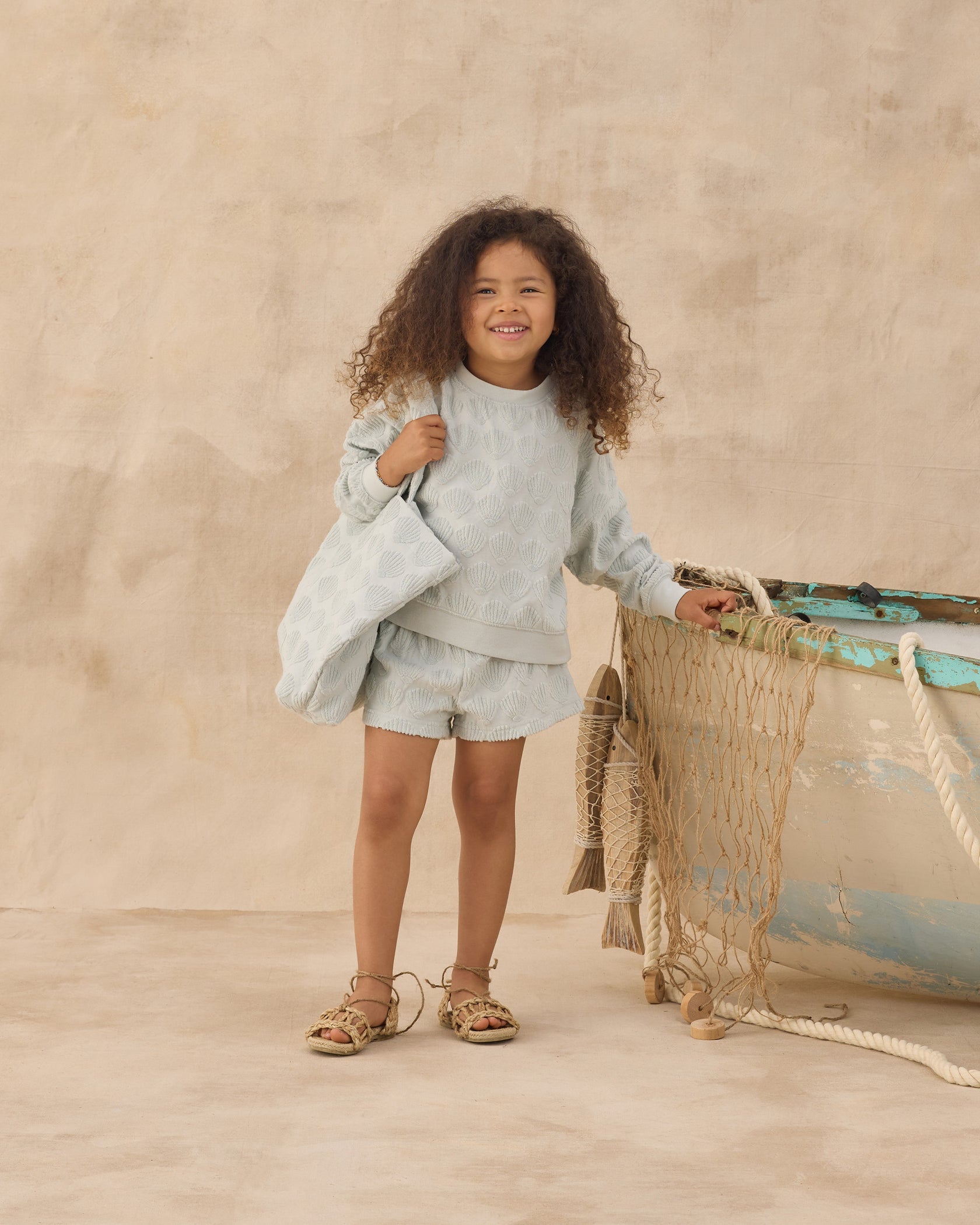 A young child with curly hair stands indoors, dressed in a light blue textured top, matching shorts, and sandals, holding the Rylee & Cru Beach Bag—an everyday accessory—beside a weathered boat adorned with netting.