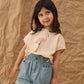 A young girl with dark hair stands in front of a crumpled brown paper background, wearing a flamingo print t-shirt paired with the Girls' Alba Denim Short by Rylee & Cru, featuring patch pockets and an elastic waistband.