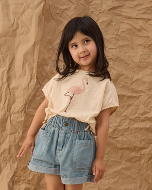 A young girl with dark hair stands in front of a crumpled brown paper background, wearing a flamingo print t-shirt paired with the Girls' Alba Denim Short by Rylee & Cru, featuring patch pockets and an elastic waistband.