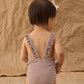 A toddler stands facing away, wearing the Rylee & Cru Girls' Positano One-Piece and a small bow headband. The background is textured brown paper.