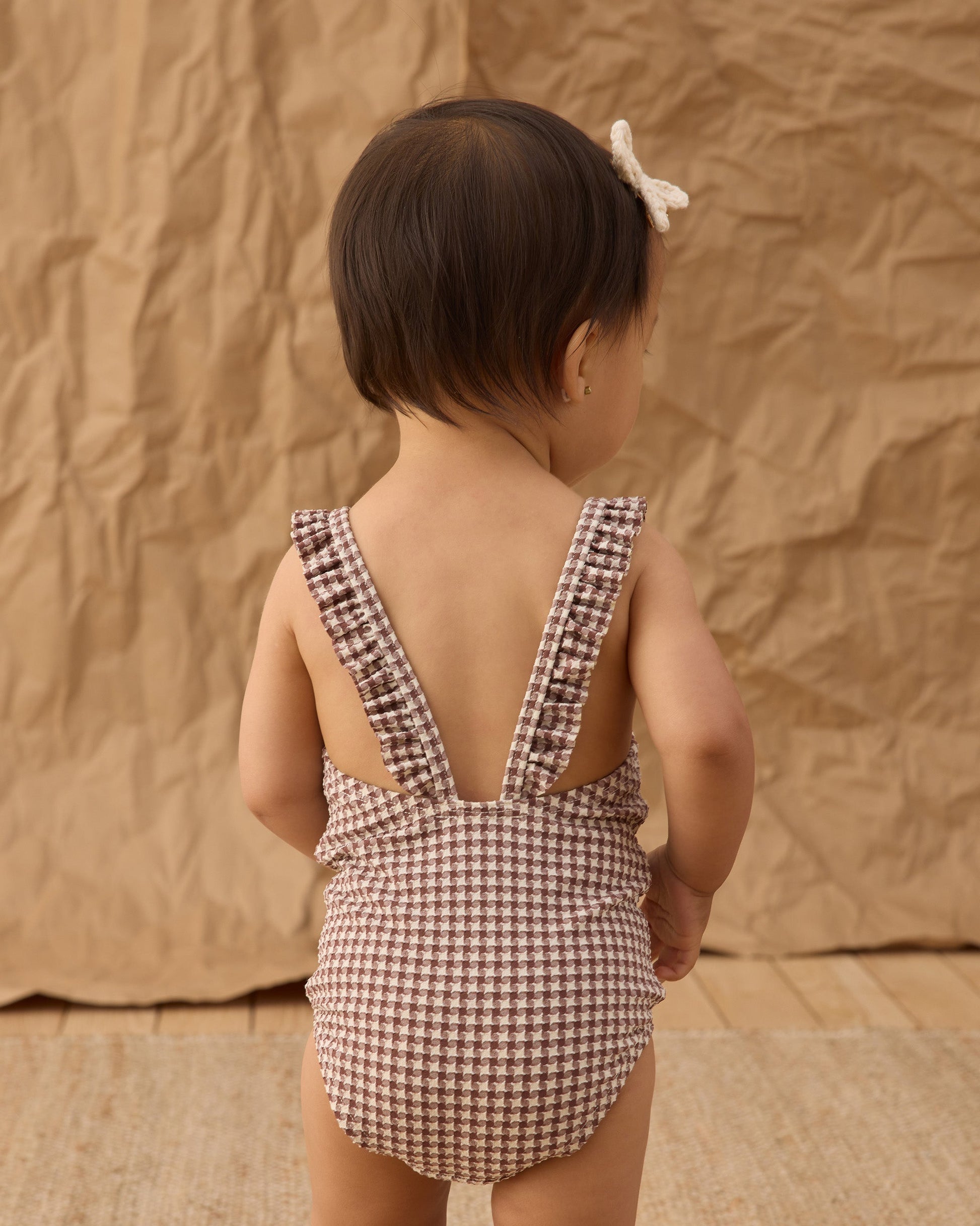 A toddler stands facing away, wearing the Rylee & Cru Girls' Positano One-Piece and a small bow headband. The background is textured brown paper.