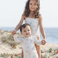A young girl and a baby boy wearing Rylee & Cru Baby Boys' Rashguard Set UPF 50+ swimwear stand on a sandy beach with green plants and the ocean in the background.