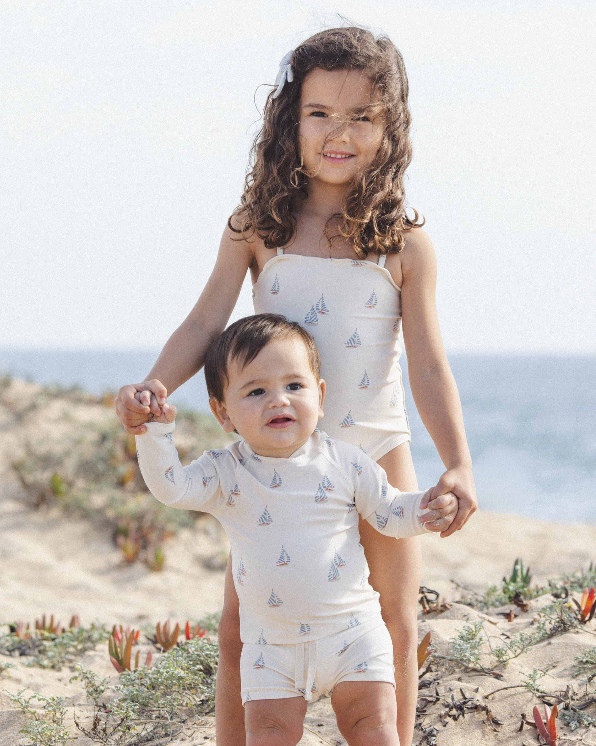 A young girl and a baby boy wearing Rylee & Cru Baby Boys' Rashguard Set UPF 50+ swimwear stand on a sandy beach with green plants and the ocean in the background.