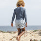 A boy with curly hair, barefoot by the ocean on sandy ground, wears a long-sleeve shirt and Rylee & Cru Baby Boys' and Boys' Swim Trunk with a sharks print.