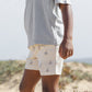 A child stands outdoors on sandy ground, wearing a light grey t-shirt and Rylee & Cru Baby Boys' and Boys' Boardshorts featuring sailboat and sun patterns. Only the lower half of their face and body are visible.