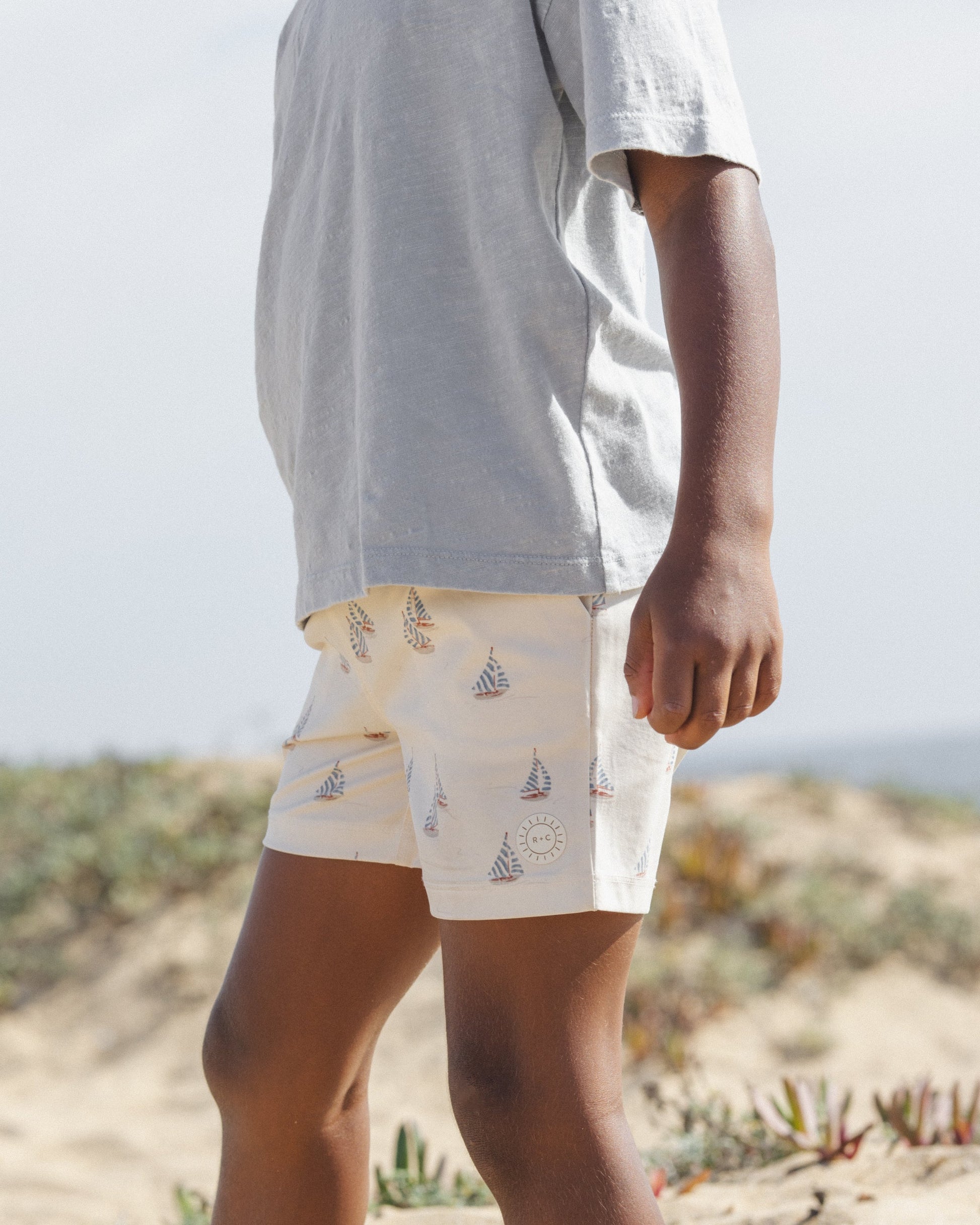 A child stands outdoors on sandy ground, wearing a light grey t-shirt and Rylee & Cru Baby Boys' and Boys' Boardshorts featuring sailboat and sun patterns. Only the lower half of their face and body are visible.