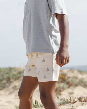 A child stands outdoors on sandy ground, wearing a light grey t-shirt and Rylee & Cru Baby Boys' and Boys' Boardshorts featuring sailboat and sun patterns. Only the lower half of their face and body are visible.