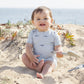 A baby in a light blue Rylee & Cru Baby Boys' Shorty One-Piece with a shark pattern sits on sandy ground near plants, holding a small white shovel, with the ocean visible in the background.