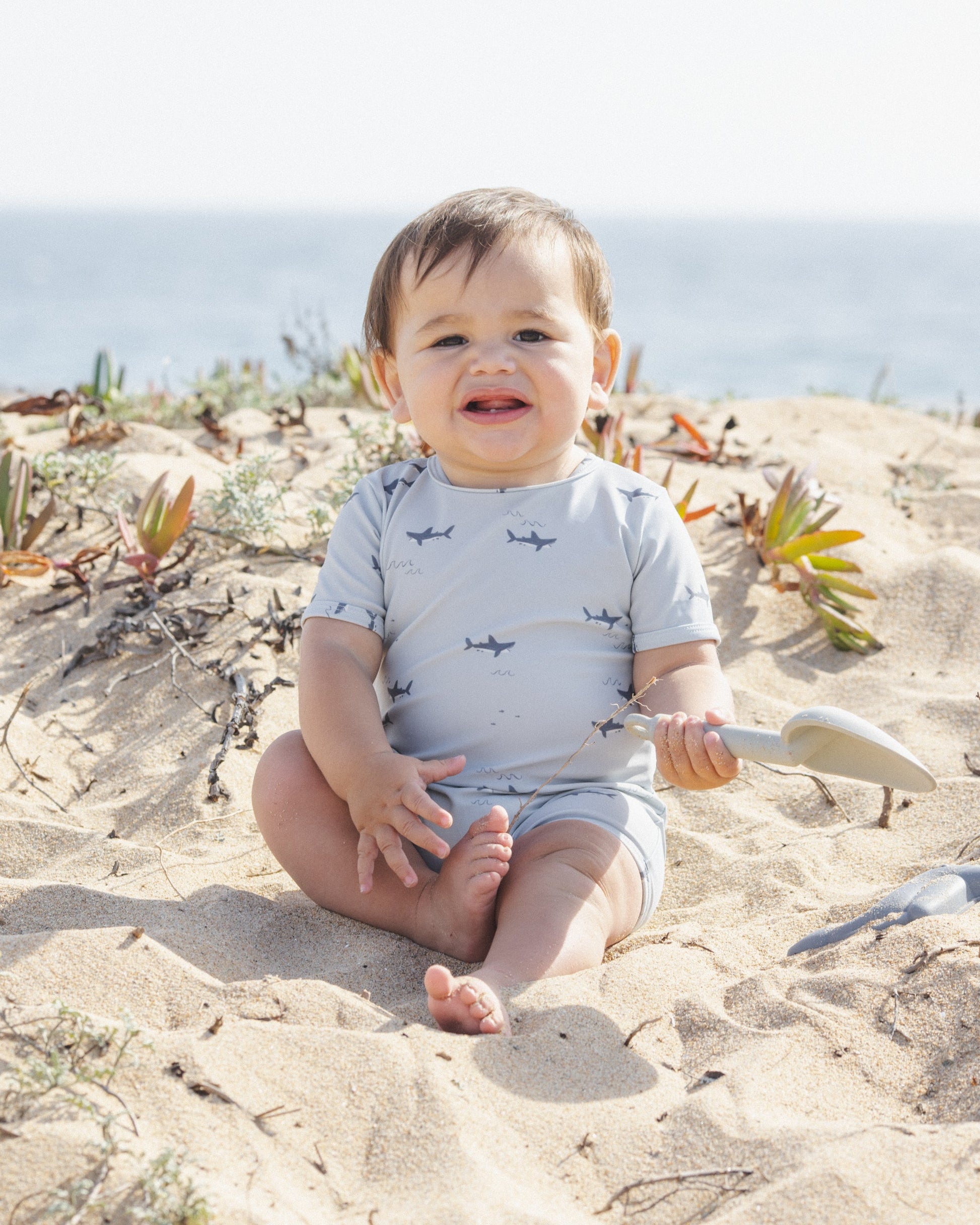 A baby in a light blue Rylee & Cru Baby Boys' Shorty One-Piece with a shark pattern sits on sandy ground near plants, holding a small white shovel, with the ocean visible in the background.