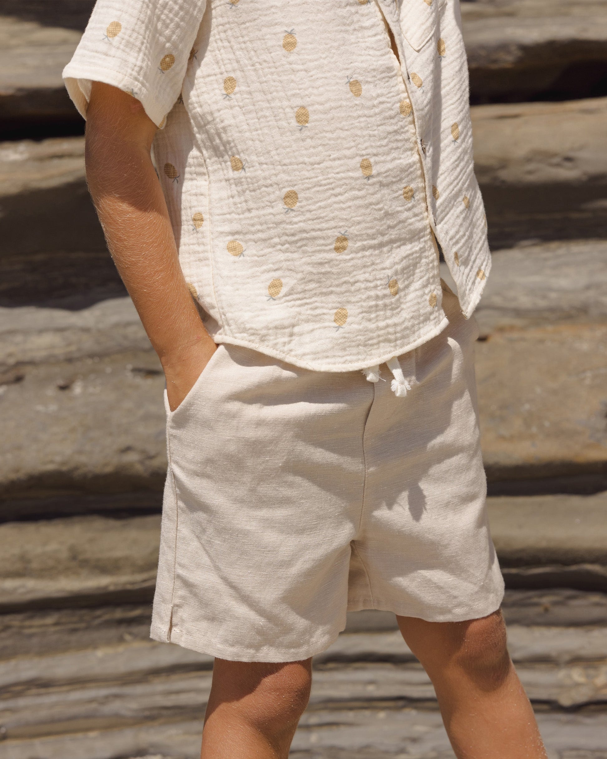A child stands hands-in-pockets, wearing a cream short-sleeve polka dot shirt and matching Rylee & Cru Baby Boys' Perry Shorts with an elastic waistband, creating a vintage look against a rocky backdrop.