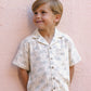 A young boy with light brown hair wears the Rylee & Cru Boys' Lapel Collar Shirt featuring a palm tree pattern as he stands smiling in front of a pale pink textured wall.