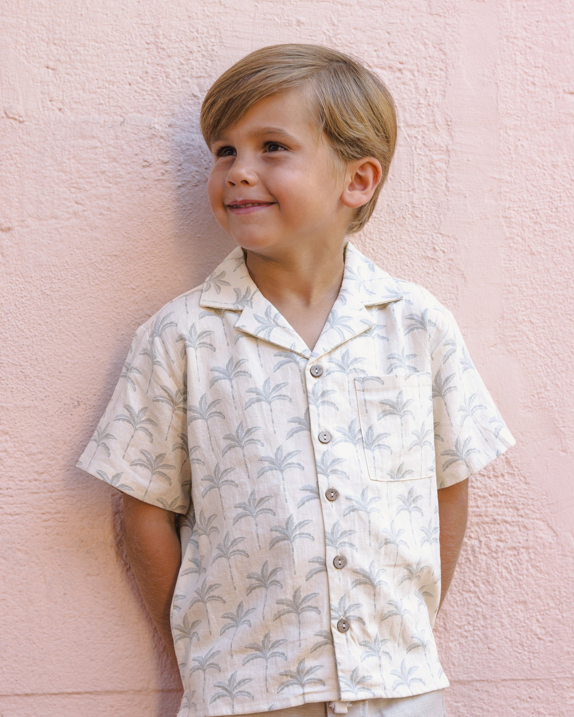A young boy with light brown hair wears the Rylee & Cru Boys' Lapel Collar Shirt featuring a palm tree pattern as he stands smiling in front of a pale pink textured wall.