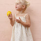 A young blonde girl in the Rylee & Cru Girls' Sahara Mini Dress with adjustable straps holds a lemon and looks aside, posing against a light pink textured wall.