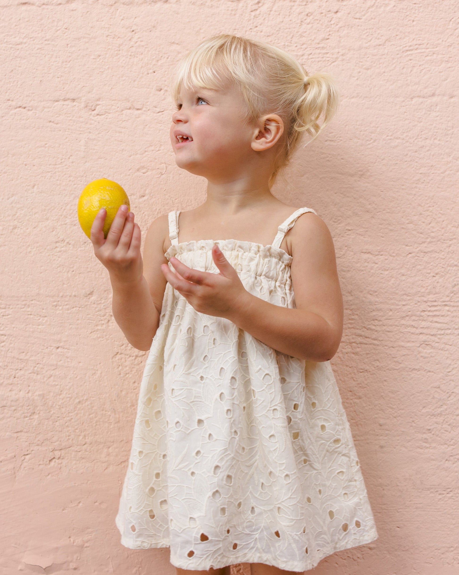 A young blonde girl in the Rylee & Cru Girls' Sahara Mini Dress with adjustable straps holds a lemon and looks aside, posing against a light pink textured wall.
