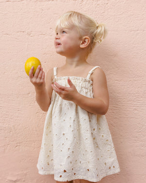 A young blonde girl in the Rylee & Cru Girls' Sahara Mini Dress with adjustable straps holds a lemon and looks aside, posing against a light pink textured wall.