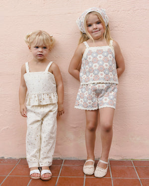 Two young children stand by a light pink wall, both wearing the Girls' Kayli Set by Rylee & Cru—light-colored, lace-trimmed summer outfits featuring a cropped tank and wide-leg pants—ideal for warm weather and paired with sandals.