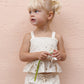 A young girl with blonde pigtails wears the Girls' Kayli Set by Rylee & Cru and holds a white flower in front of a pale pink wall—an adorable look for warm weather days.