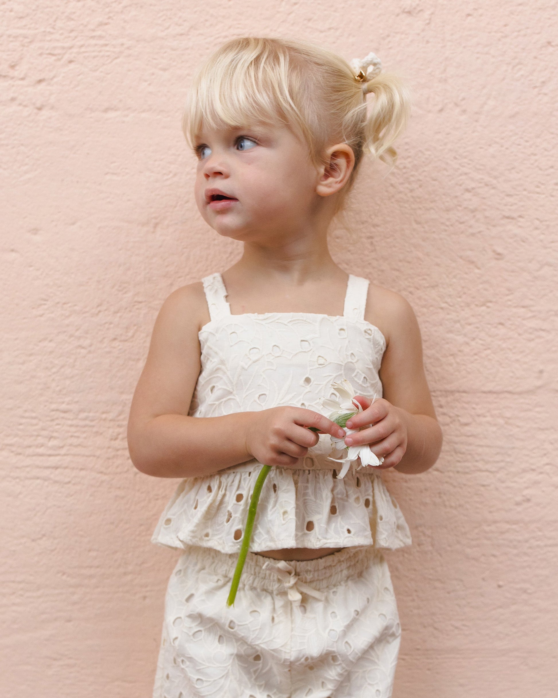 A young girl with blonde pigtails wears the Girls' Kayli Set by Rylee & Cru and holds a white flower in front of a pale pink wall—an adorable look for warm weather days.