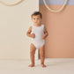 A toddler models the Rylee & Cru Baby Girls' and Girls' Moxie One-Piece in periwinkle stripes with UPF 50+ protection, barefoot on a light floor against a color-blocked backdrop, ropes on the wall, and finished with a light blue bow.