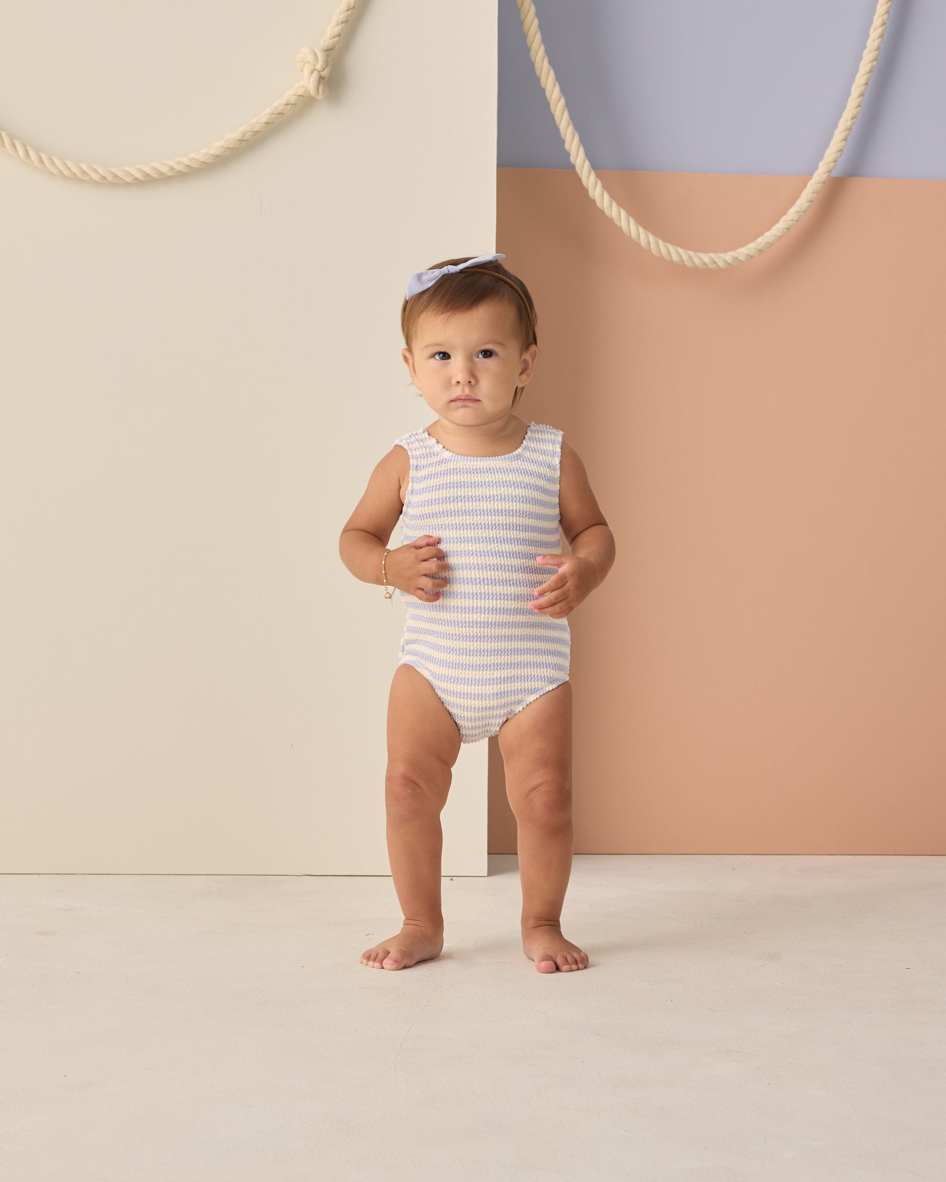 A toddler models the Rylee & Cru Baby Girls' and Girls' Moxie One-Piece in periwinkle stripes with UPF 50+ protection, barefoot on a light floor against a color-blocked backdrop, ropes on the wall, and finished with a light blue bow.