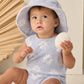 A baby in the Rylee & Cru Baby Girls' Cinch Playsuit in periwinkle daisy print sits with a white seashell, wearing a floral hat, against a dried palm leaf and light background.