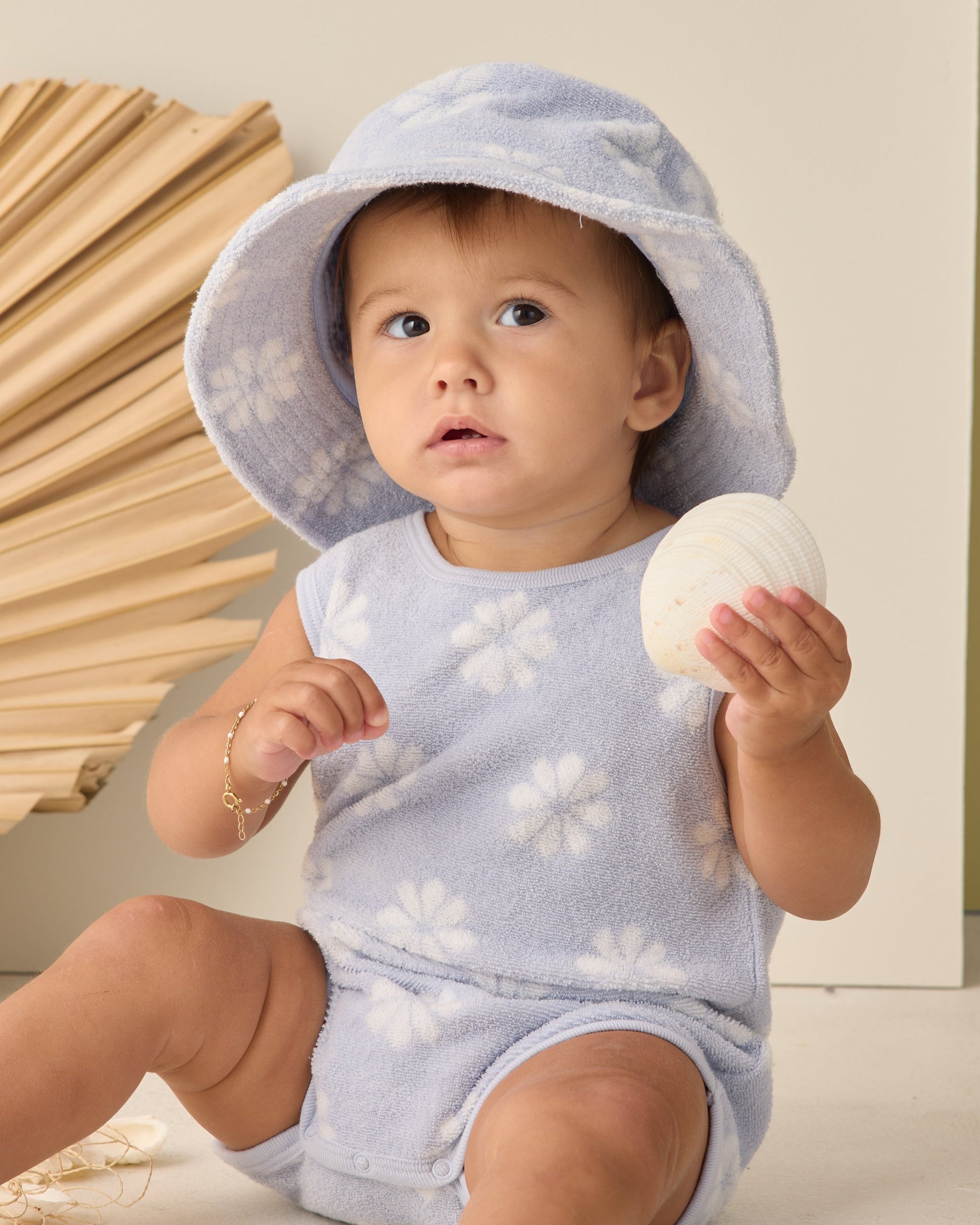 A baby in the Rylee & Cru Baby Girls' Cinch Playsuit in periwinkle daisy print sits with a white seashell, wearing a floral hat, against a dried palm leaf and light background.