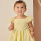 A young child in a yellow Rylee & Cru dress and a Rylee & Cru Baby Bow Headband stands indoors, smiling and looking slightly aside.