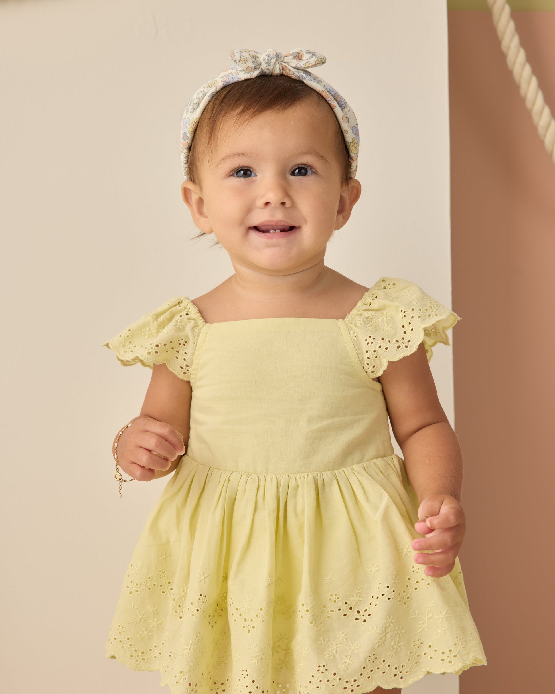 A young child in a yellow Rylee & Cru dress and a Rylee & Cru Baby Bow Headband stands indoors, smiling and looking slightly aside.
