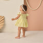 A toddler wears the Rylee & Cru Baby Girls' Remi Romper in yellow with eyelet detail and a headband, standing in a minimal studio with pastel walls and rope accents.