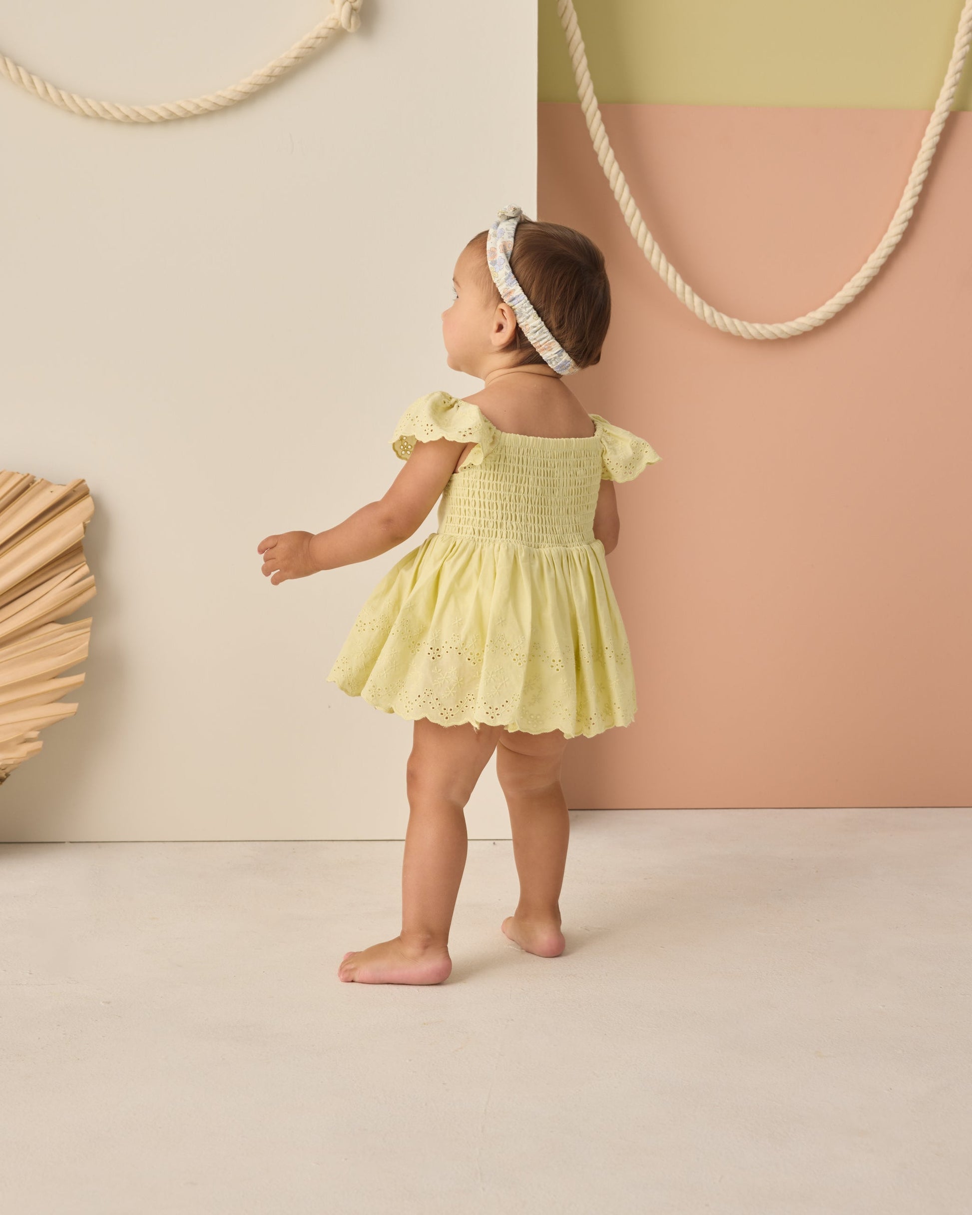 A toddler wears the Rylee & Cru Baby Girls' Remi Romper in yellow with eyelet detail and a headband, standing in a minimal studio with pastel walls and rope accents.
