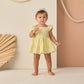 A toddler in a comfy Rylee & Cru Baby Girls' Remi Romper with eyelet detail and a headband stands barefoot indoors against a pastel backdrop with decorative rope and paper fans.