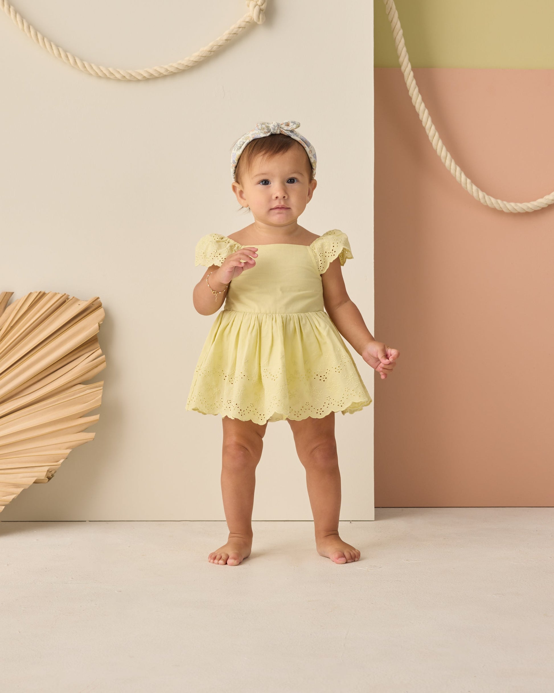 A toddler in a comfy Rylee & Cru Baby Girls' Remi Romper with eyelet detail and a headband stands barefoot indoors against a pastel backdrop with decorative rope and paper fans.