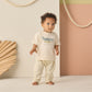 A toddler stands indoors wearing a cream t-shirt with a crocodile graphic and Rylee & Cru's Baby Rowan Pant featuring stripes and an elastic waistband, against a backdrop of decorative ropes and geometric wall panels.