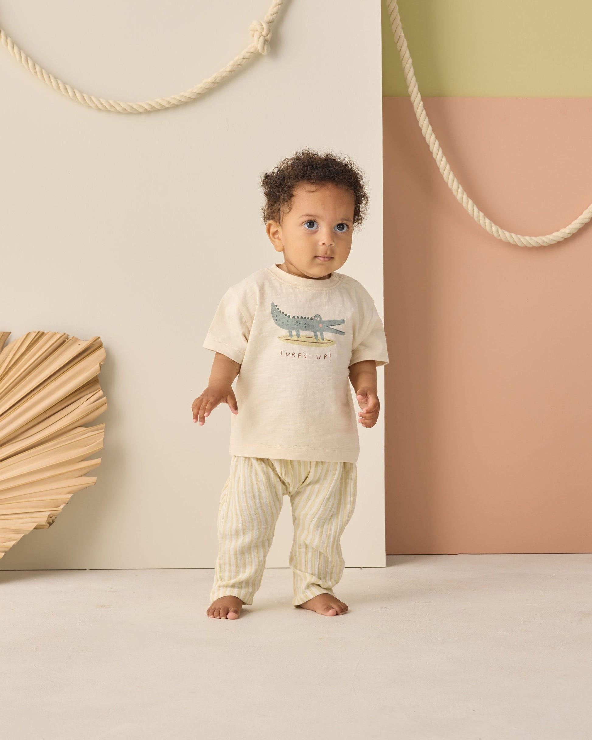 A toddler stands indoors wearing a cream t-shirt with a crocodile graphic and Rylee & Cru's Baby Rowan Pant featuring stripes and an elastic waistband, against a backdrop of decorative ropes and geometric wall panels.