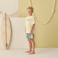 Young boy stands barefoot indoors, wearing a striped yellow and white "SURF" tee and Rylee & Cru Baby Boys' and Boys' Paneled Boardshorts, with a surfboard and rope decor in the background.