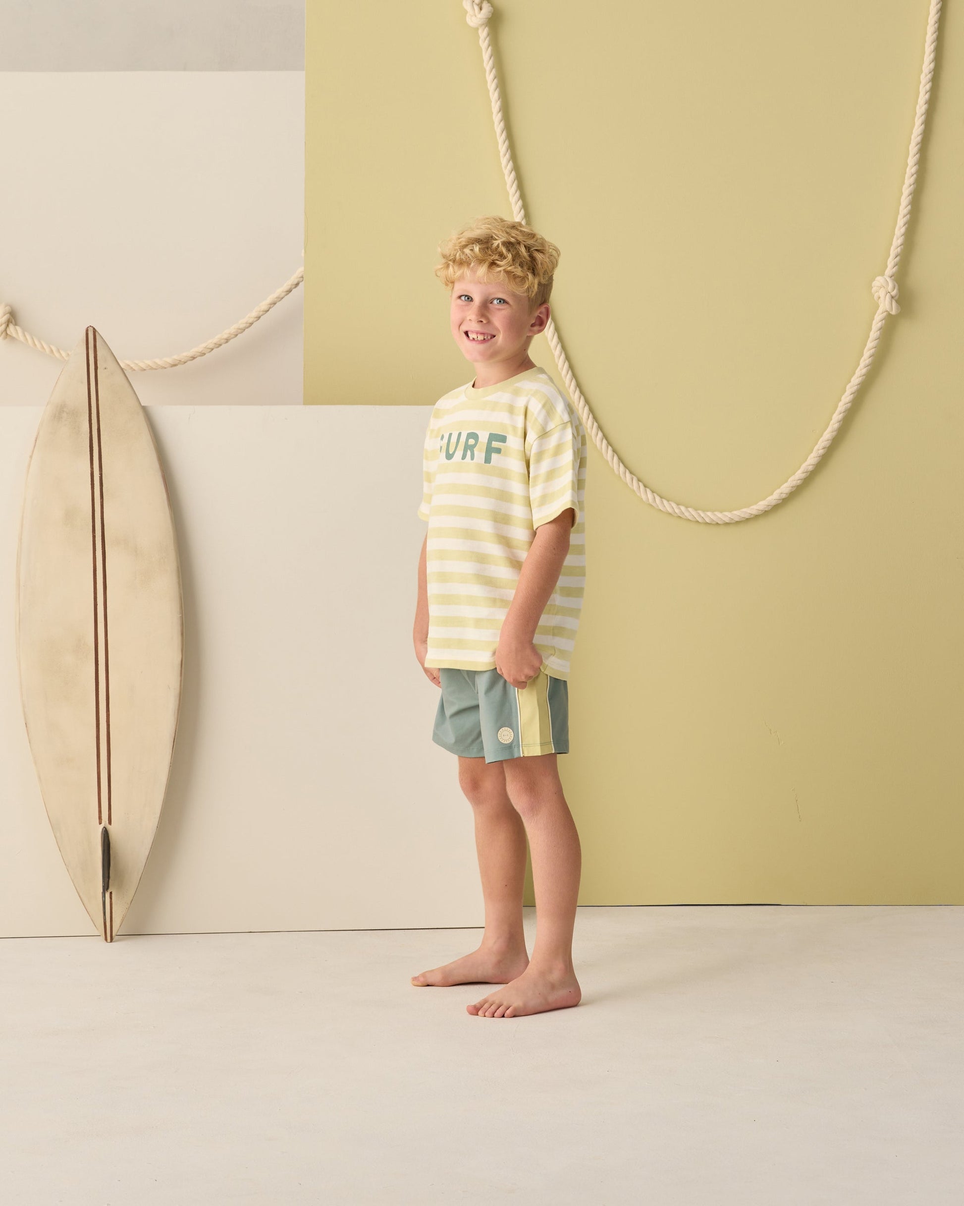 Young boy stands barefoot indoors, wearing a striped yellow and white "SURF" tee and Rylee & Cru Baby Boys' and Boys' Paneled Boardshorts, with a surfboard and rope decor in the background.