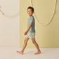 A young boy smiles barefoot indoors in Rylee & Cru Baby Boys' and Boys' Swim Trunk, featuring striped elastic drawstrings and a mesh liner, set against a minimalist backdrop with decorative ropes.