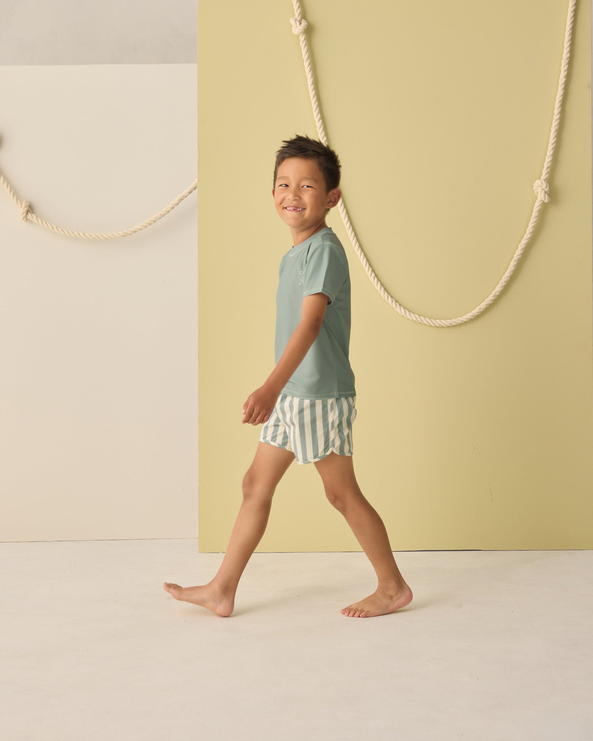 A young boy smiles barefoot indoors in Rylee & Cru Baby Boys' and Boys' Swim Trunk, featuring striped elastic drawstrings and a mesh liner, set against a minimalist backdrop with decorative ropes.