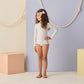 A young girl with curly hair stands barefoot on a white floor, wearing the Rylee & Cru Baby Girls' and Girls' Rashguard One-Piece, in front of pastel-colored blocks and a rope on the wall.