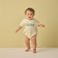 A baby stands on a light floor by a beige wall, wearing the Rylee & Cru Baby Boys' Relaxed Bubble Romper—yellow and white striped with "SURF" printed on the front.