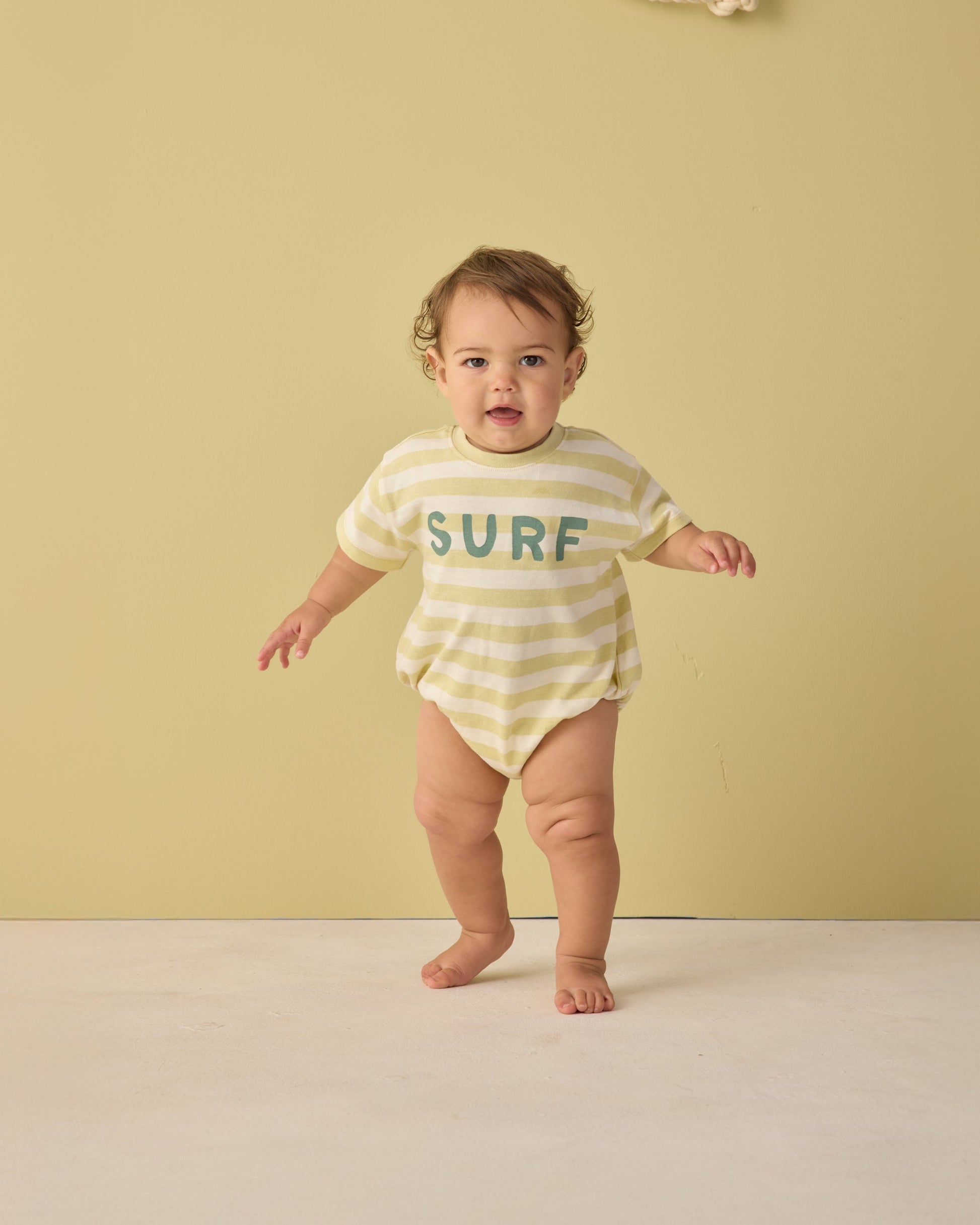 A baby stands on a light floor by a beige wall, wearing the Rylee & Cru Baby Boys' Relaxed Bubble Romper—yellow and white striped with "SURF" printed on the front.
