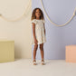 Indoors, a young girl wears the Girls' Lucia Dress by Rylee & Cru, a charming blue floral summer dress, paired with white sandals; pastel blocks and rope decorations adorn the background.